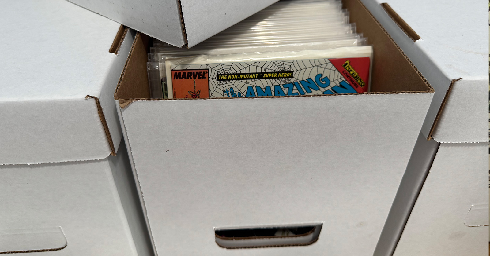 Stacked white cardboard comic storage boxes, one open to reveal bagged-and-boarded comics including an Amazing Spider-Man issue.