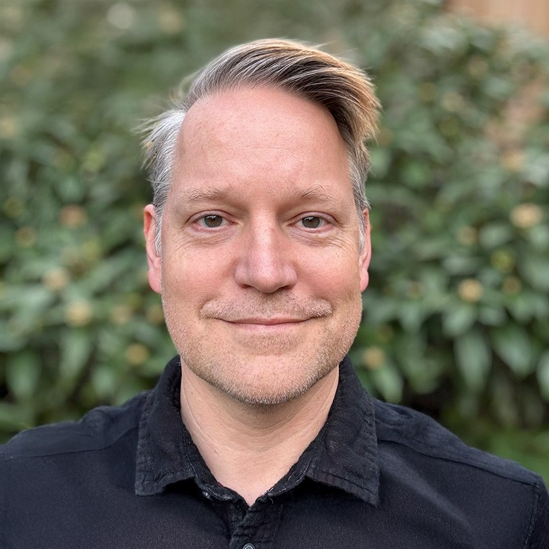 Portrait of Adam Kinney, founder of Comic Scry — short hair, dark shirt, photographed in front of green foliage.