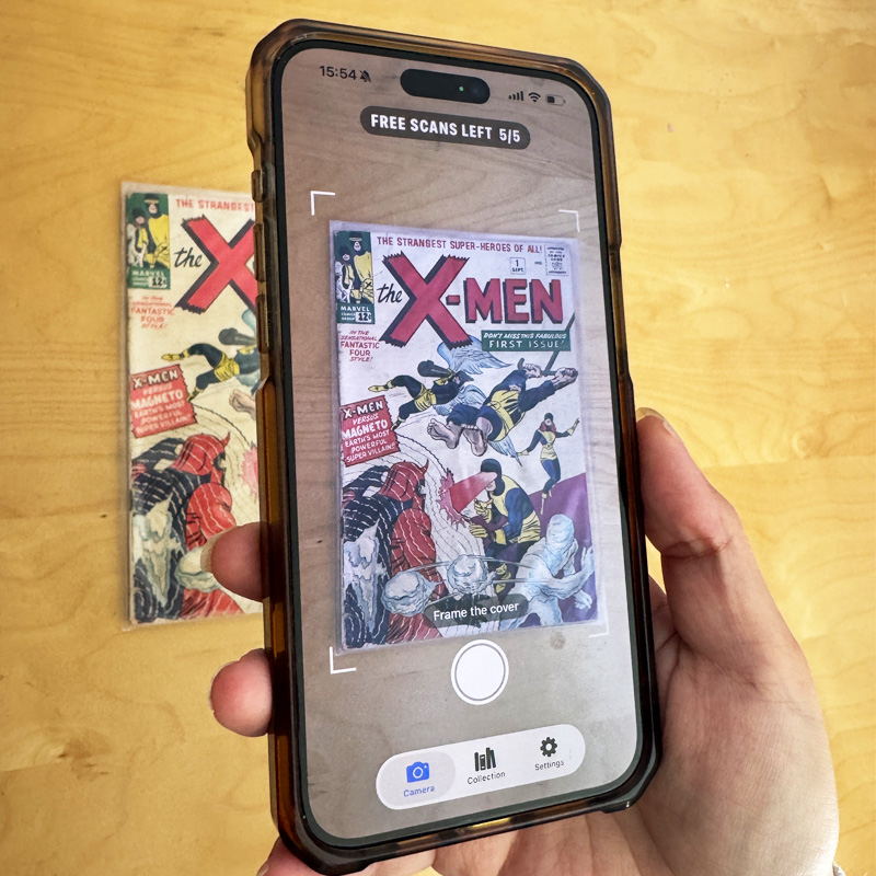 A hand holding a phone running Comic Scry, framing an X-Men #1 comic book cover laid on a wood table. The on-screen reticle says "Frame the cover."